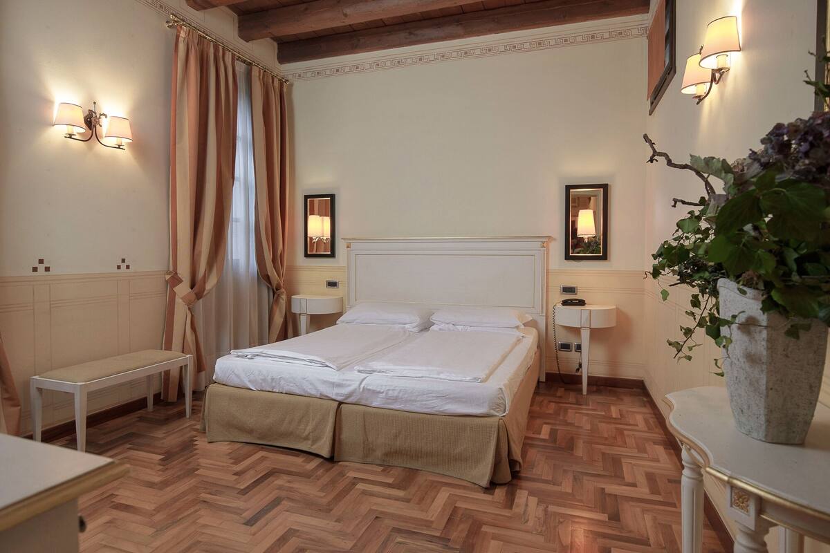 Ein Hotelzimmer mit einem großen Bett, zwei Nachttischen, einer Nachttischlampe, einem Spiegel, einem kleinen Tisch und einer Topfpflanze.