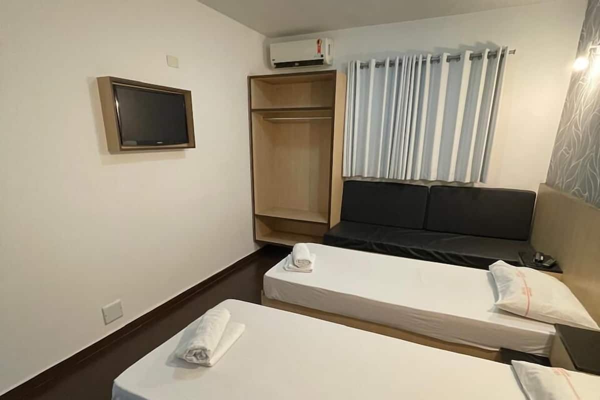 Quarto de hotel com duas camas de solteiro, televisão na parede, guarda-roupa e janela com cortinas.