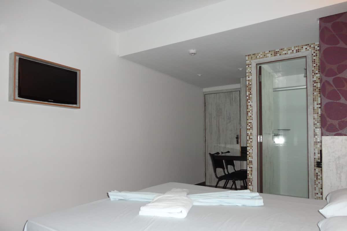 Quarto de hotel com cama, televisão na parede e banheiro visível através de uma porta de vidro.