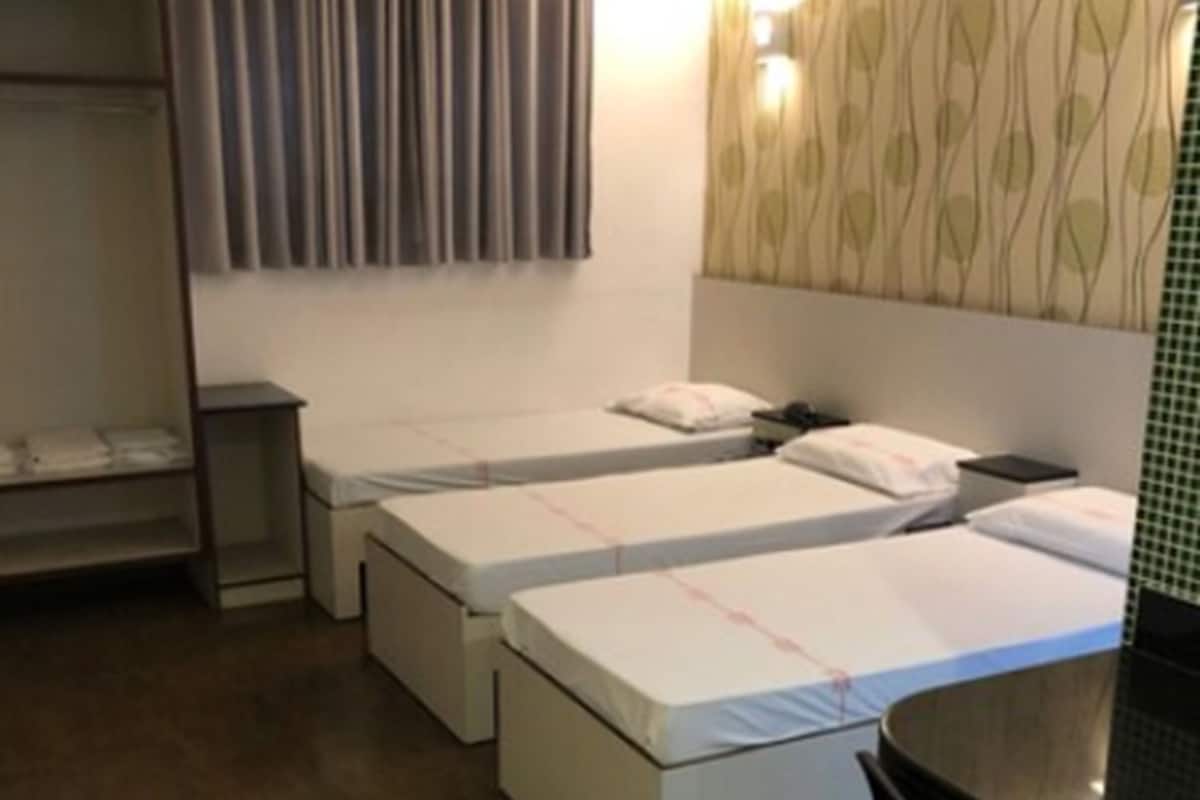Quarto de hotel com duas camas de solteiro, uma escrivaninha e uma cadeira. Há um guarda-roupa e uma prateleira com objetos.