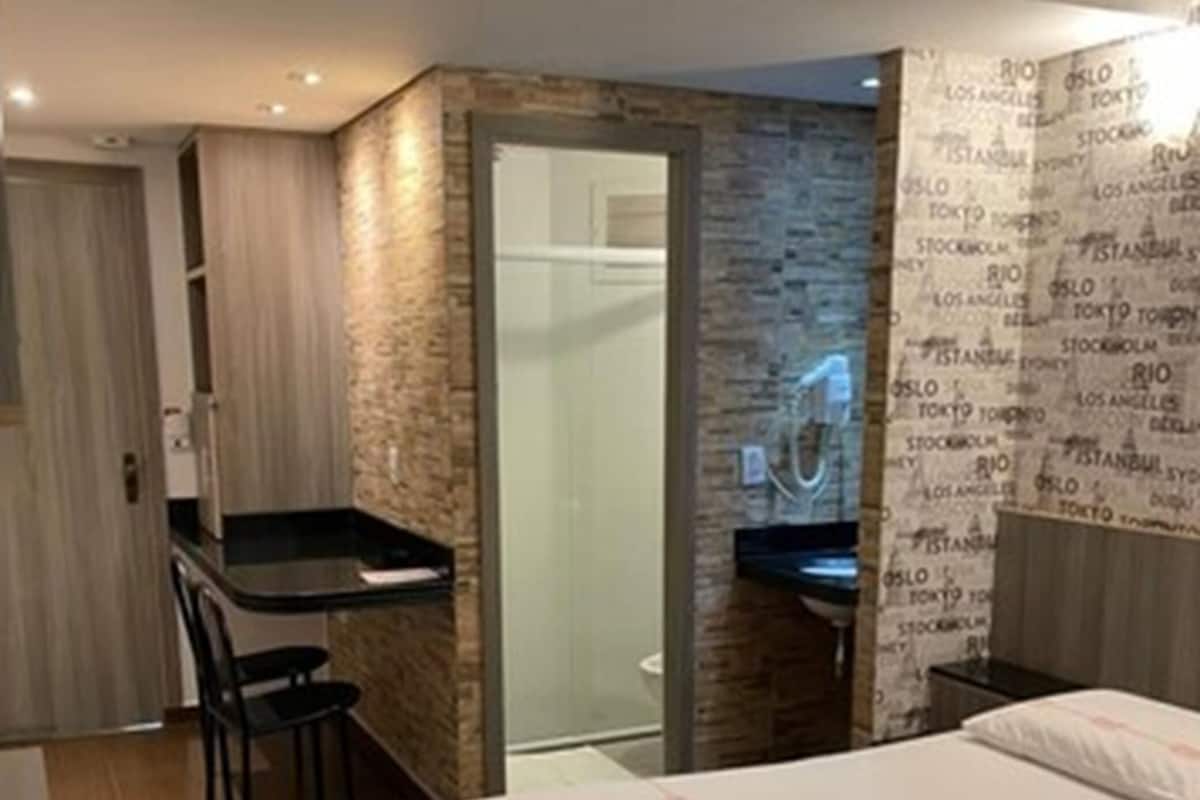 Quarto de hotel compacto com cama, escrivaninha e área de chuveiro.