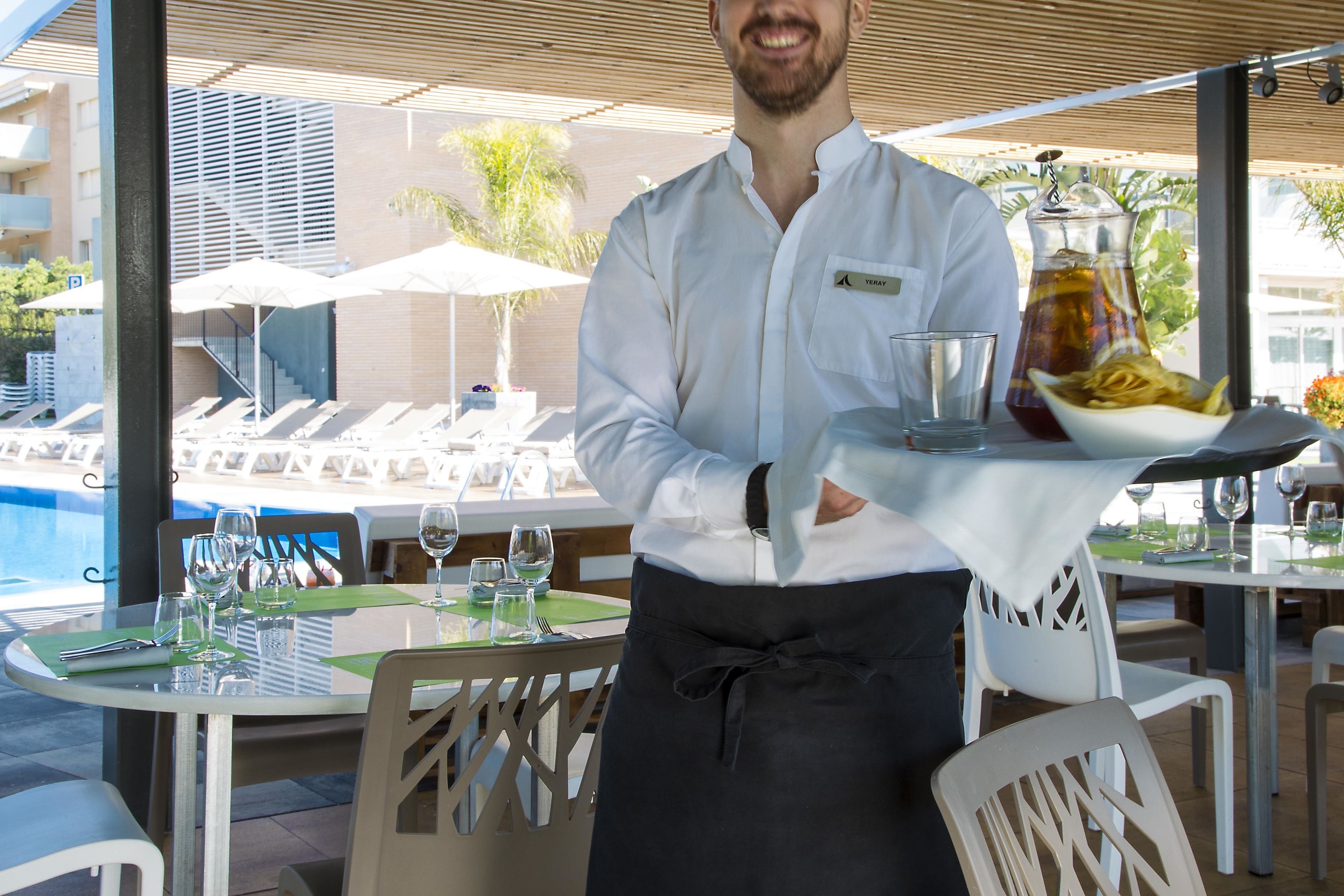 Un camarero sirve una bandeja con una jarra, vasos y un plato de comida en una zona de comedor al aire libre con vistas a la piscina.