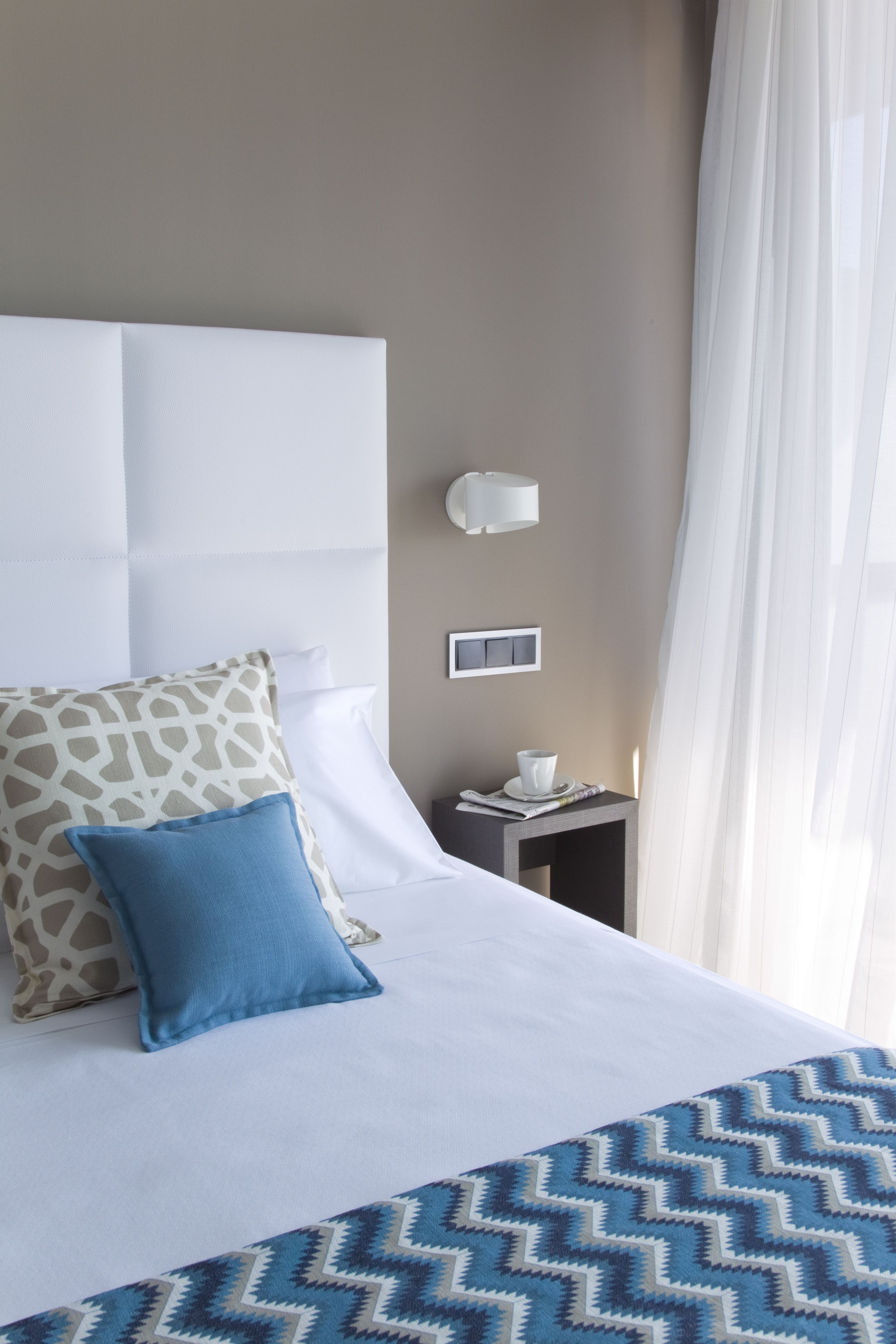 A hotel room with a large bed, a nightstand, a wall-mounted lamp, and a window with sheer curtains.