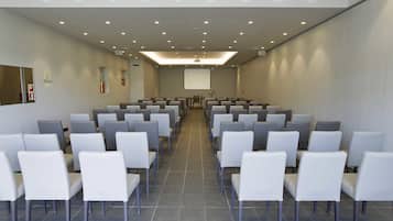 Un salón de conferencias con filas de sillas blancas, una pantalla de proyector y suelo de baldosas.