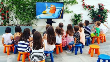 Children seated on colorful stools watching a television screen outdoors.
