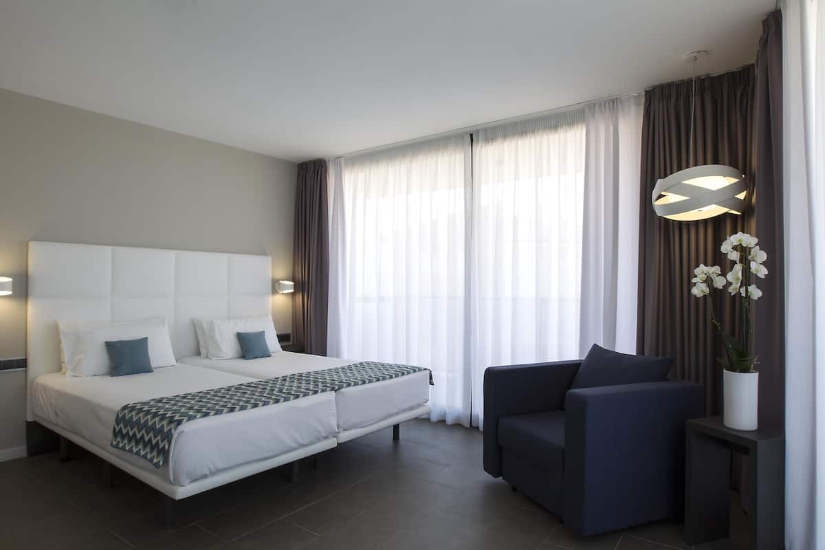 Quarto de hotel com uma cama grande, duas mesas de cabeceira, um sofá, um vaso de planta e uma janela ampla com cortinas.