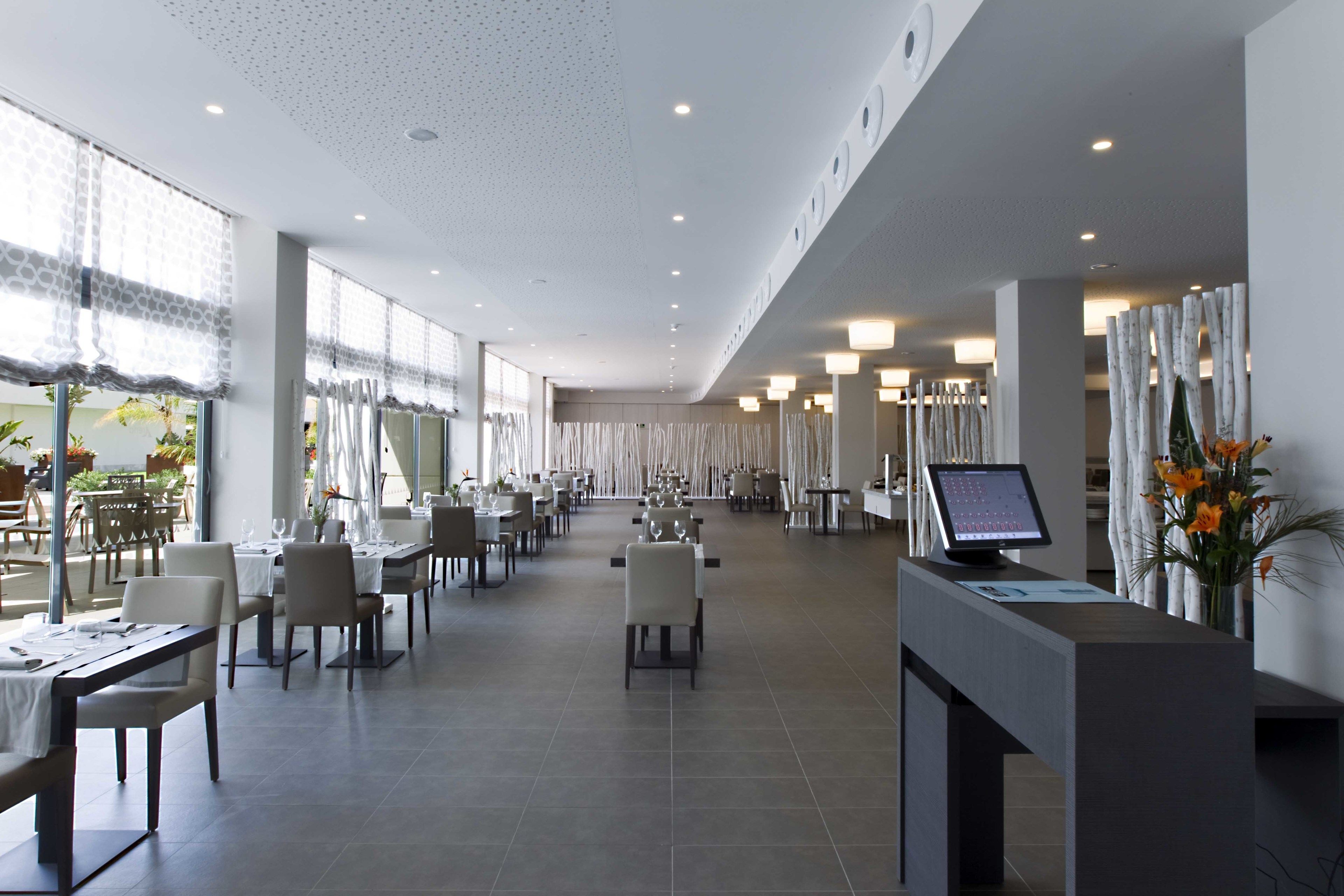 A spacious hotel dining area with multiple tables and chairs, a reception desk, and a large window providing natural light.
