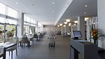 A spacious hotel dining area with multiple tables and chairs, a reception desk, and a large window providing natural light.