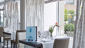 Un restaurant amb una decoració moderna, que presenta una taula llarga amb seients, un suport per al menú i una vista de l'exterior a través de grans finestres.