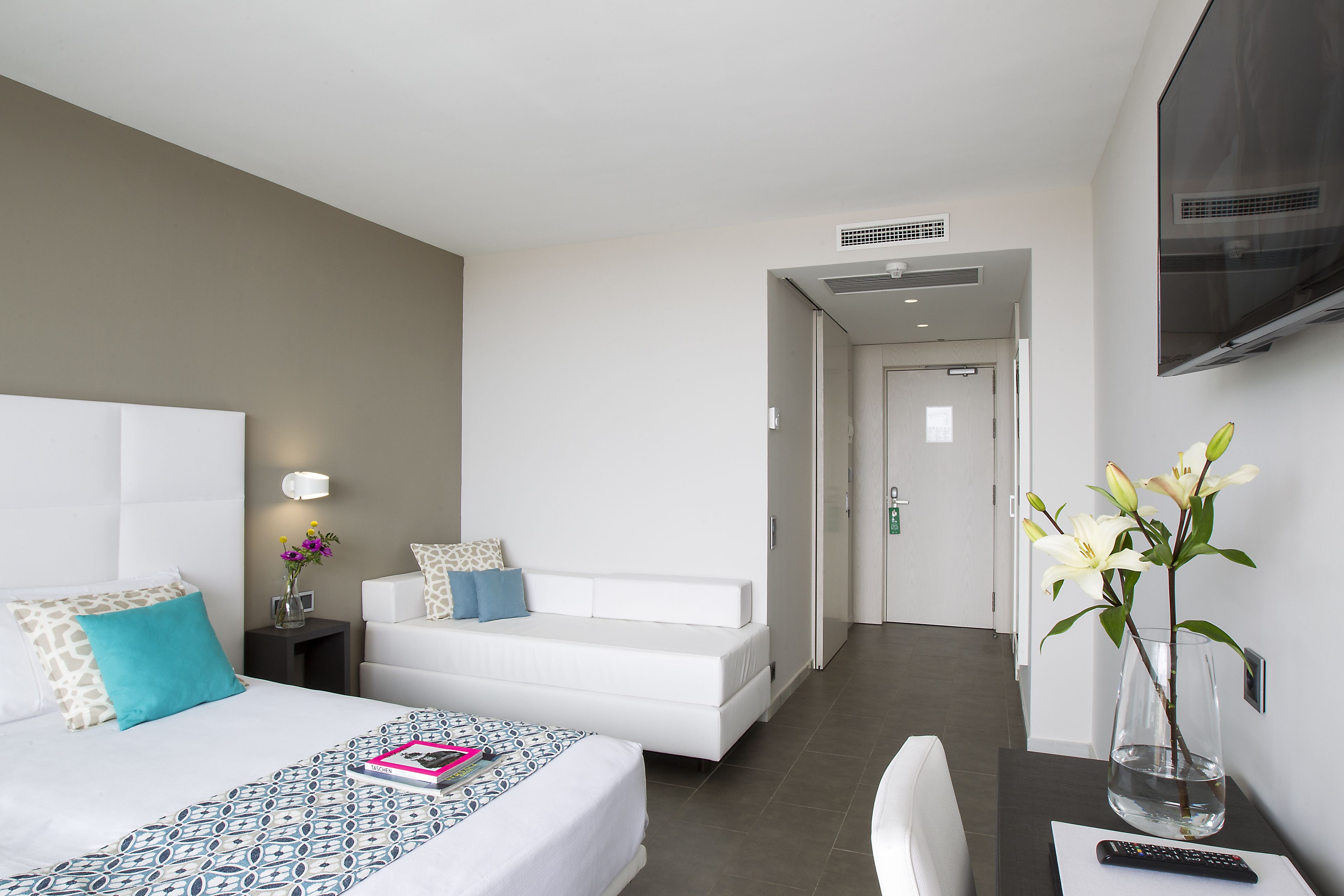 Una habitación de hotel moderna con cama, sofá blanco, escritorio con un florero y televisor montado en la pared.
