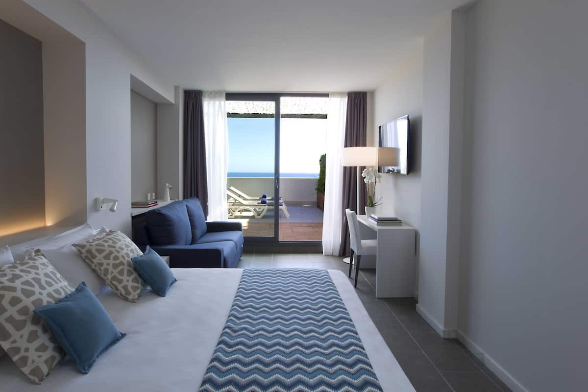 Quarto de hotel com cama, escrivaninha, cadeira, televisão e varanda com vista para o mar.