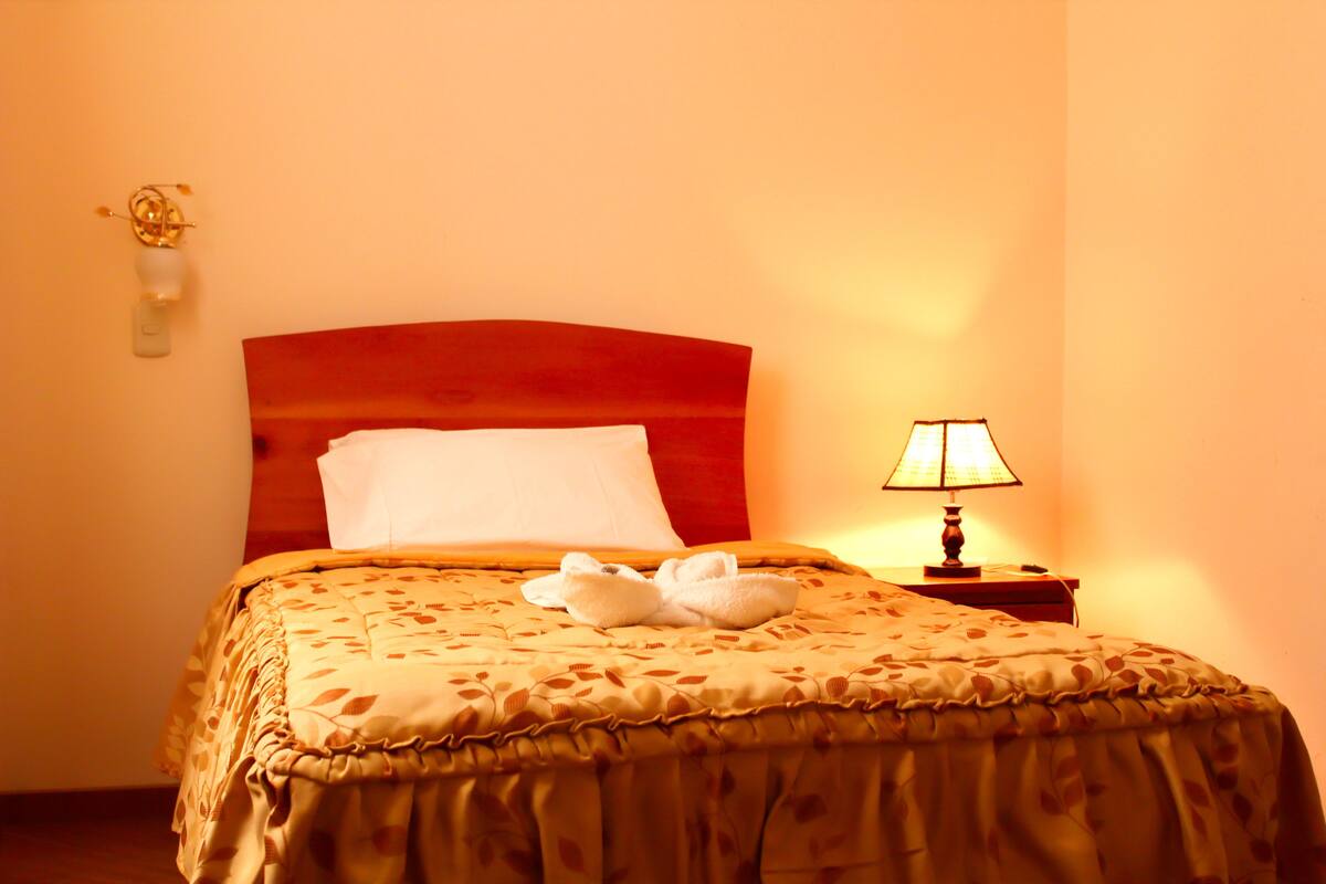 A hotel room with a bed, a nightstand, a lamp, and a wall-mounted light fixture.