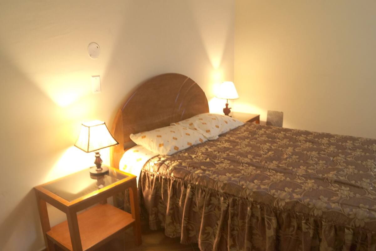 A bedroom with a wooden headboard, two bedside tables with lamps, a bed with a floral bedspread, and a nightstand with a lamp.