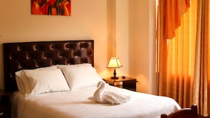 A neatly made bed with white linens, a brown tufted headboard, a white pillow, and a folded towel on the bed. There is a nightstand with a lamp and a painting on the wall above the bed. - Hotel Conquistador (Andahuaylas)