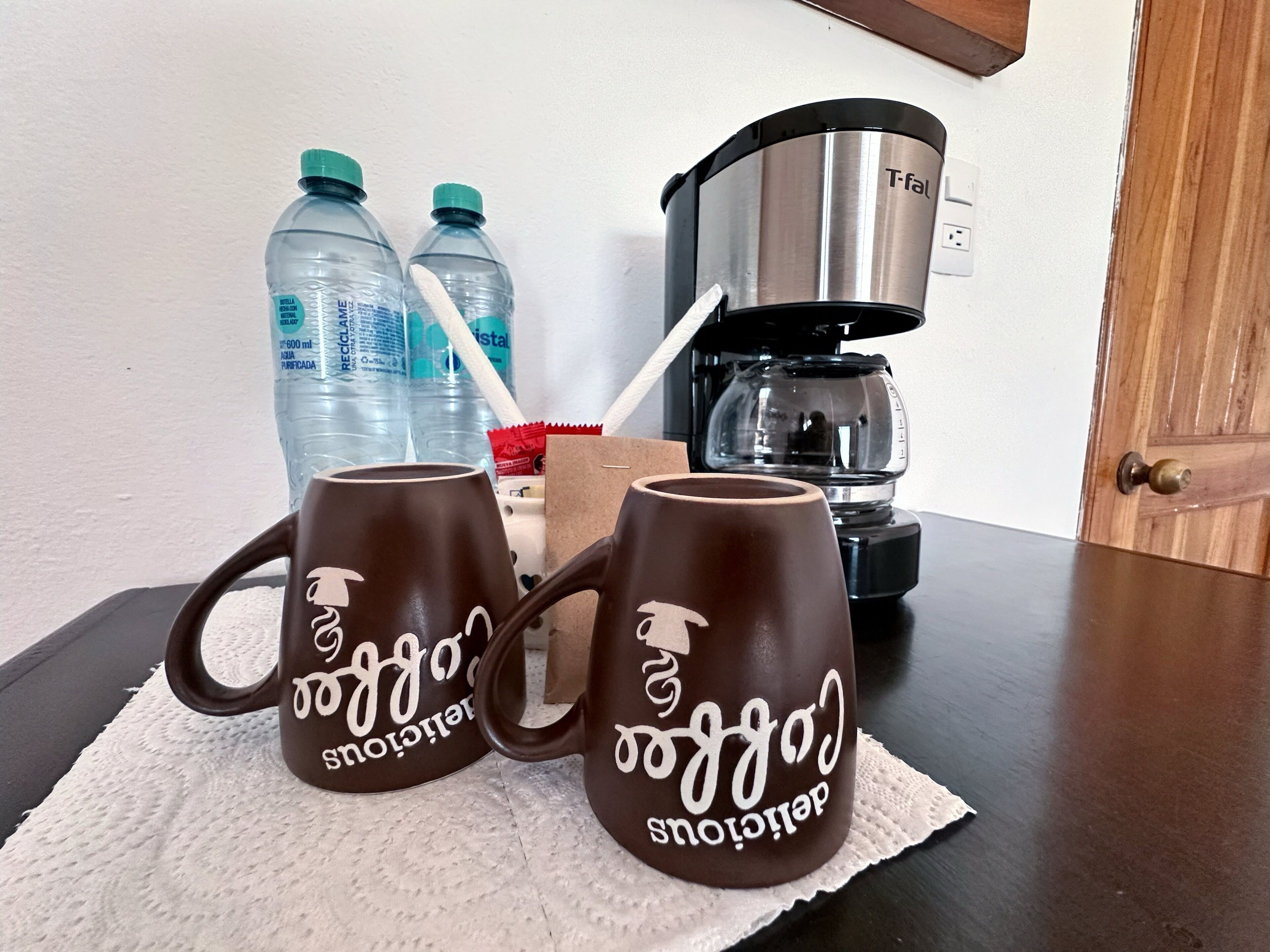 Two brown mugs with white text, a coffee maker, and two water bottles on a table.