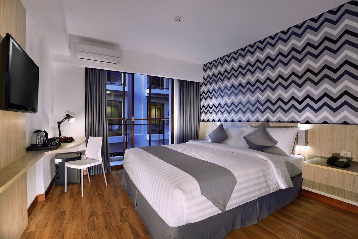 A modern hotel room with a large bed, a desk with a chair, a flat-screen TV, and a view of the building's interior through the windows.
