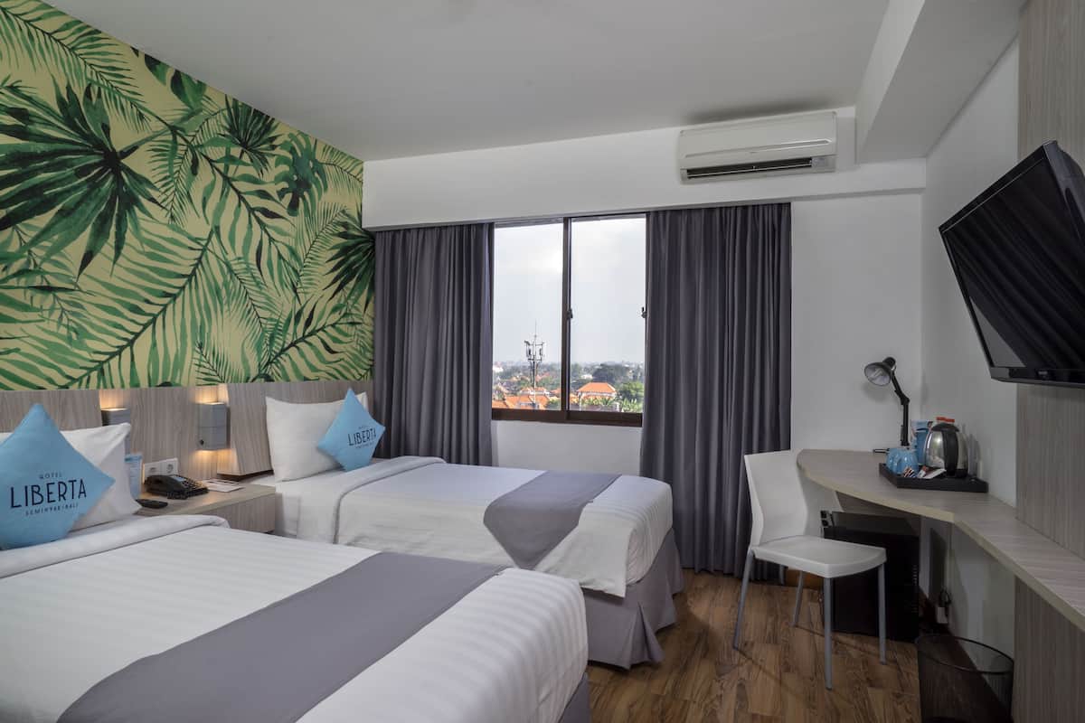 A hotel room with two beds, a desk, a chair, a TV, and a large window with curtains.
