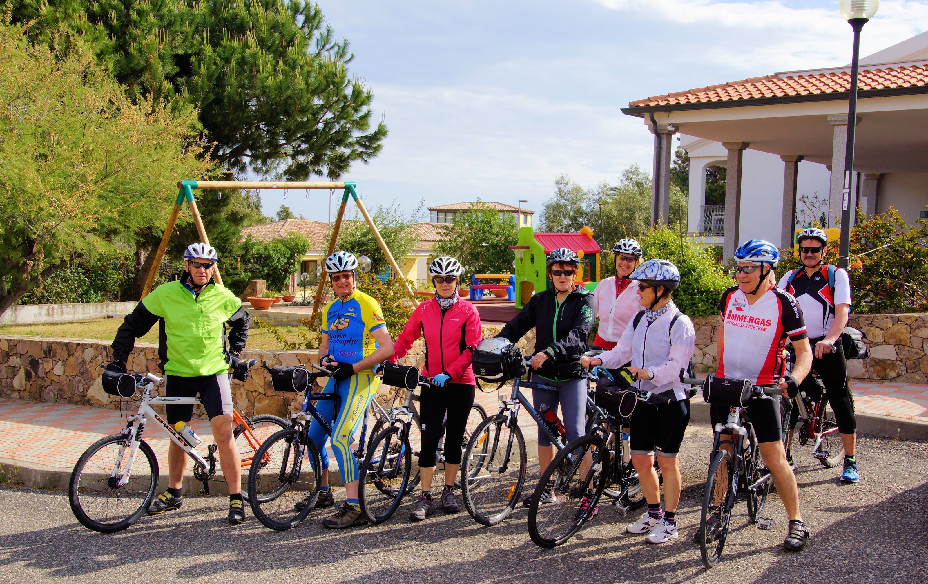 A group of cyclists standing with their bicycles in an outdoor setting with a stone wall and greenery in the background.