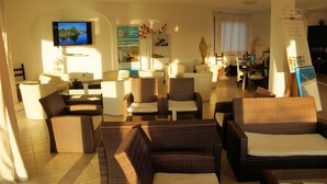 A hotel lobby with wicker furniture, a flat-screen TV, a whiteboard with notes, and a sign for a nearby hotel. - Hotel Tartaruga Bianca (Valledoria)