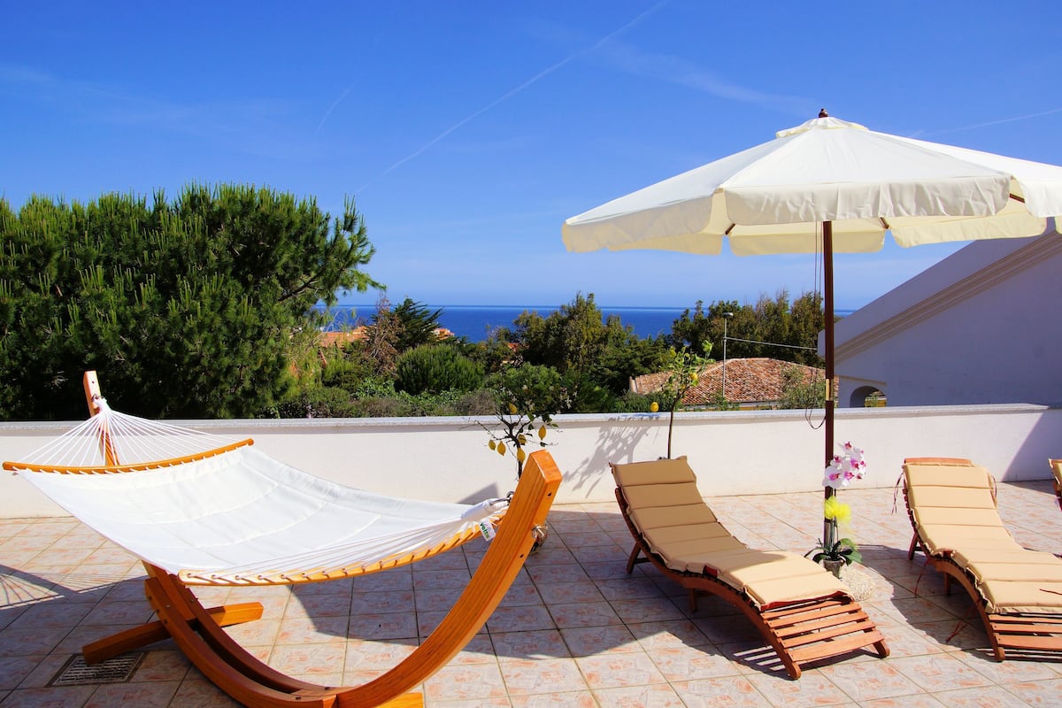 A hammock, lounge chairs, and an umbrella on a terrace with a view of the sea.