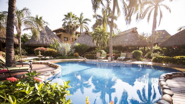 A pool surrounded by palm trees and thatched-roof structures, with lounge chairs and a well-maintained garden. - Present Moment Retreat (Troncones)