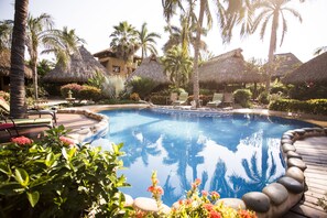 A pool surrounded by palm trees and thatched-roof structures, with lounge chairs and a well-maintained garden.
