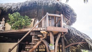A thatched-roof structure with wooden stairs leading up to a platform, surrounded by greenery and rocks. - Present Moment Retreat (Troncones)