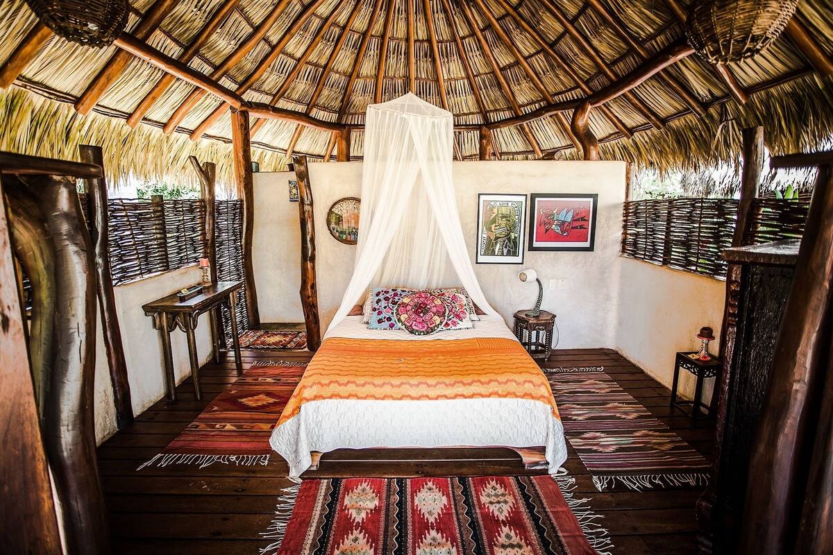 Una habitación con techo de paja, una cama con dosel blanco y alfombras estampadas.
