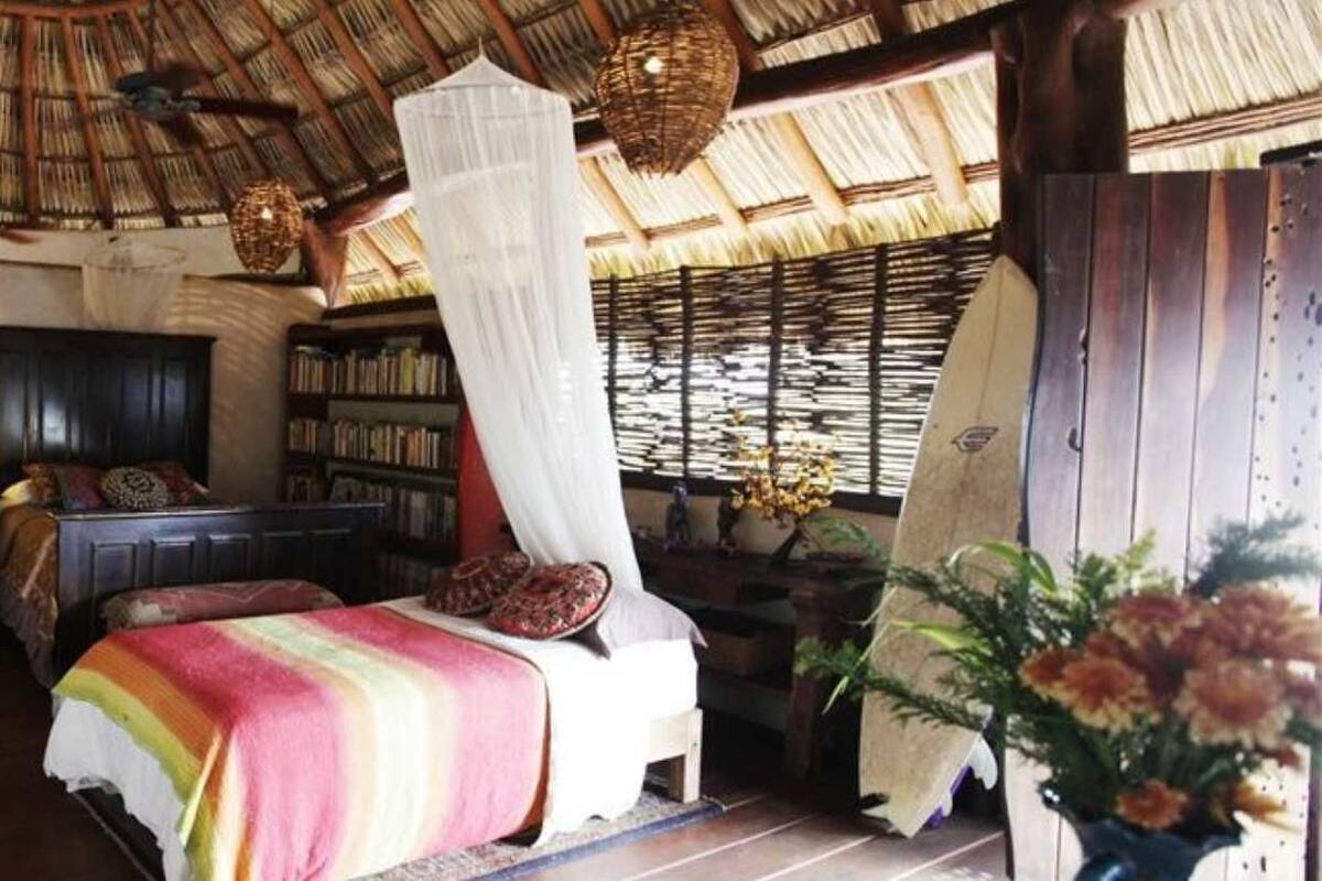 Una habitación con techo de paja, muebles de madera, una cama con colcha a rayas coloridas y una estantería.