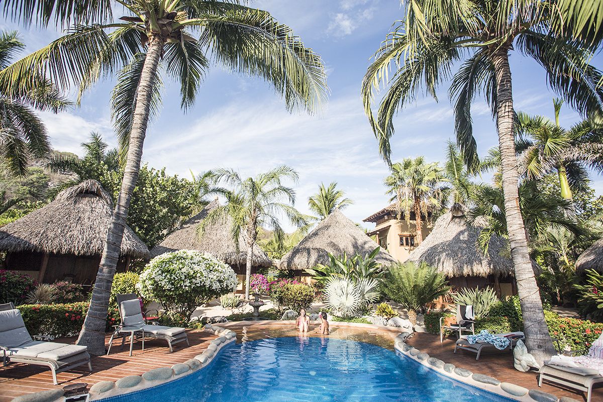 A pool area with lounge chairs, palm trees, and thatched-roof structures.