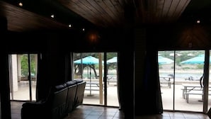 Ein Zimmer mit dunkler Holzbalkendecke, einem schwarzen Ledersofa und Blick auf eine Terrasse mit Sonnenschirmen und Tischen.