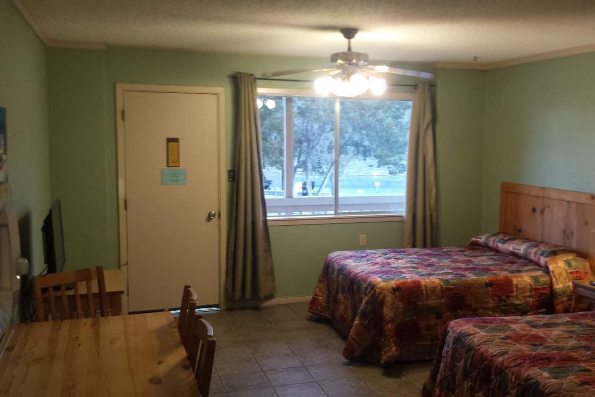 A room with two beds, a dining table, chairs, a ceiling fan, a window with curtains, and a lamp.