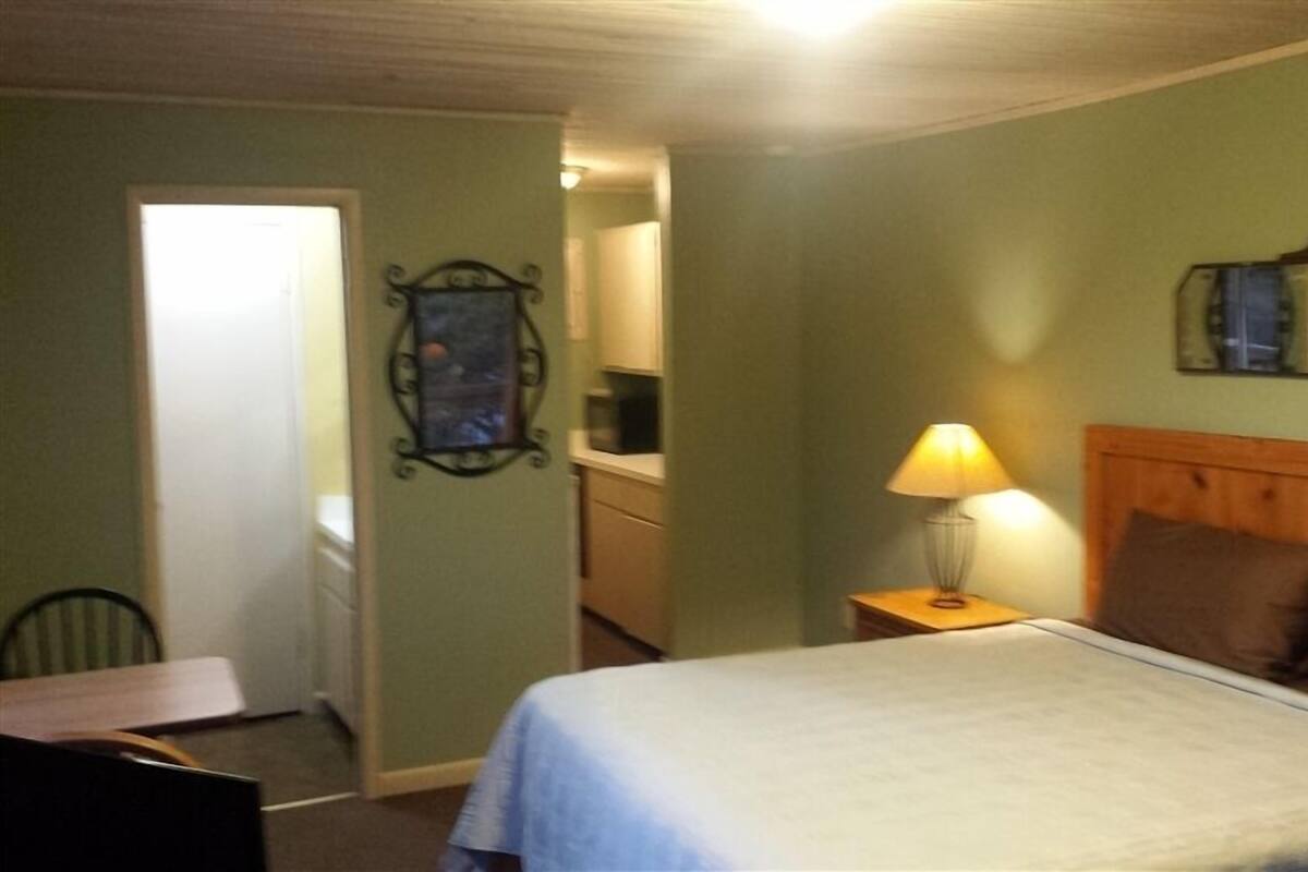 A bedroom with a bed, a nightstand with a lamp, a wooden headboard, a mirror, and a door leading to a bathroom.
