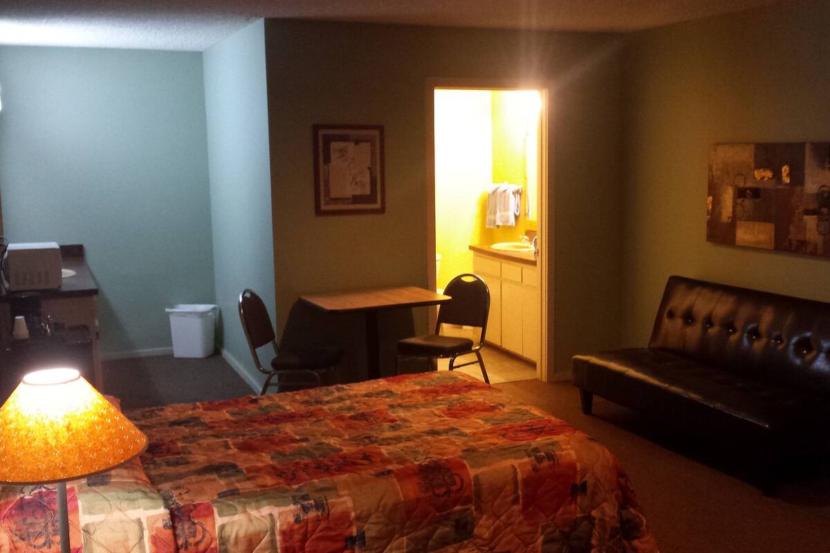 A hotel room with a bed, a desk, a chair, a couch, a lamp, and a bathroom visible through an open door.