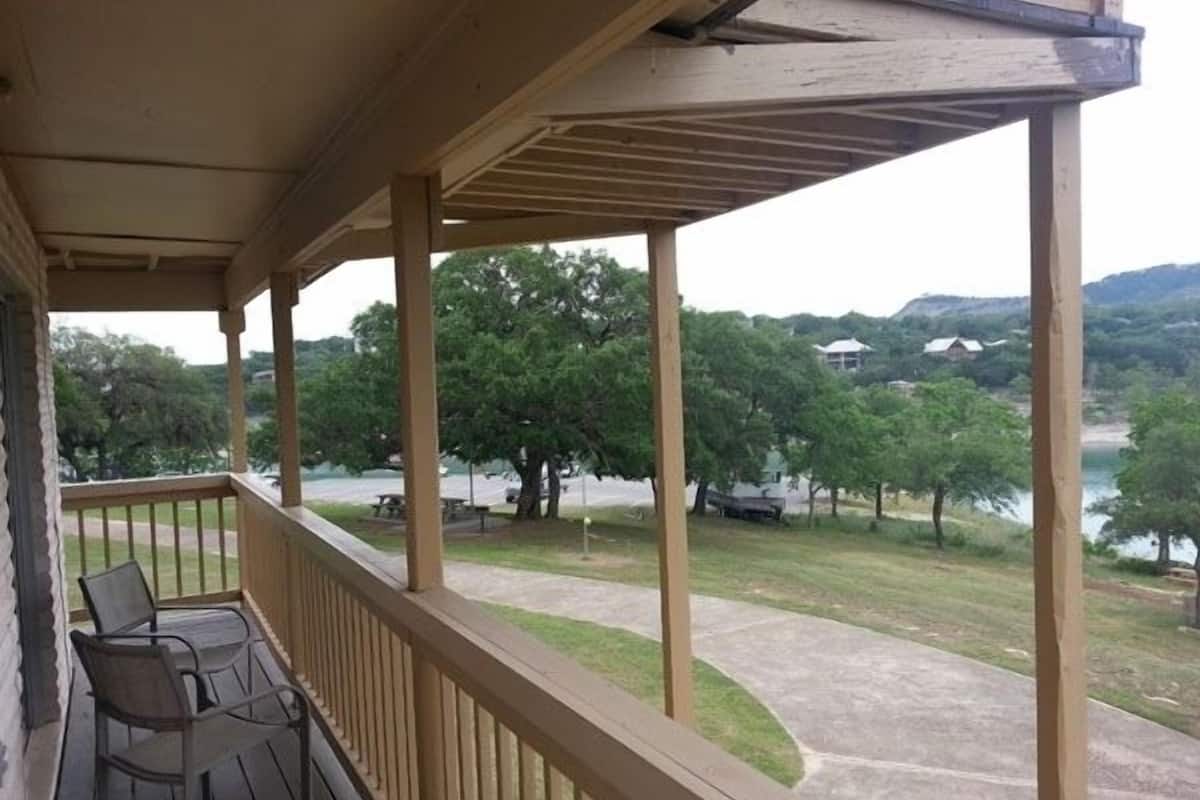 A covered porch with wooden railings and two chairs, overlooking a grassy area with trees and a body of water.