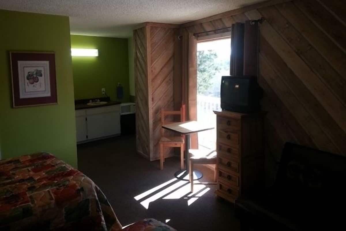 A room with a bed, a small table, a TV on a wooden cabinet, a window with curtains, and a green wall.