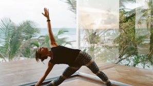 Une personne effectue une posture de yoga sur un tapis dans une pièce dotée d’une grande fenêtre donnant sur une plage et des palmiers.