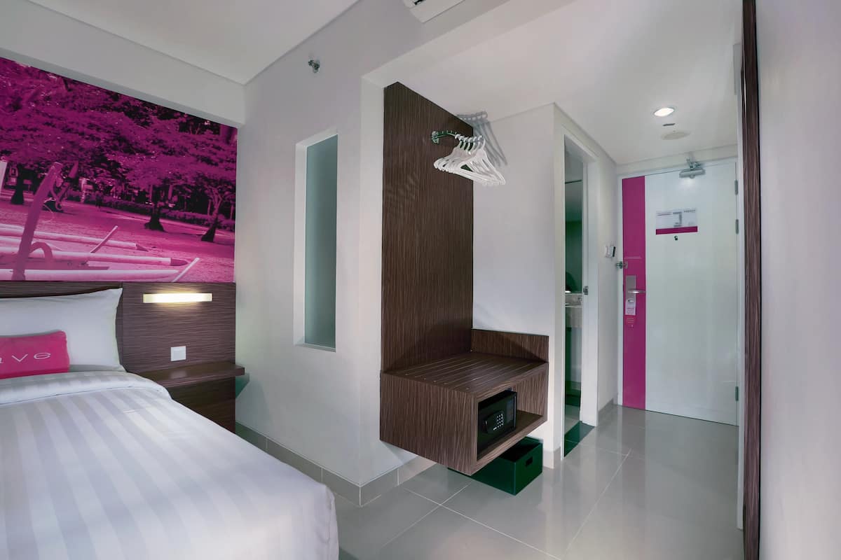A modern hotel room with a bed, a built-in TV, a wooden shelf, and a bathroom visible through an open door.