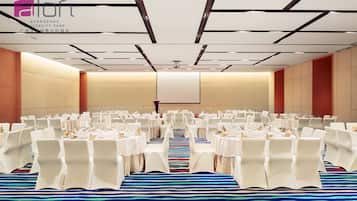 Un salón de conferencias con filas de sillas blancas y mesas, una pantalla grande al frente y una alfombra estampada.