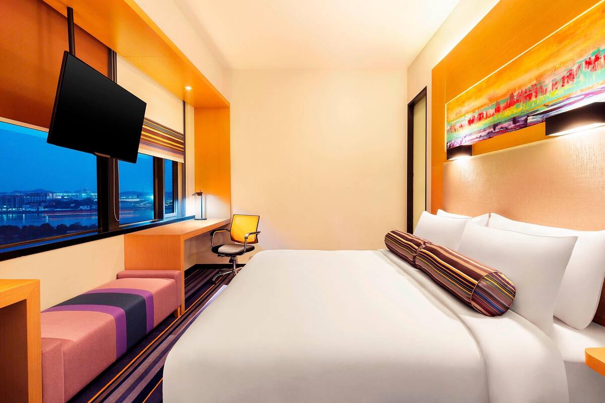 Une chambre d’hôtel moderne dotée d’un grand lit, d’un téléviseur à écran plat fixé au mur, d’un bureau avec une chaise et d’une fenêtre donnant sur la ville.