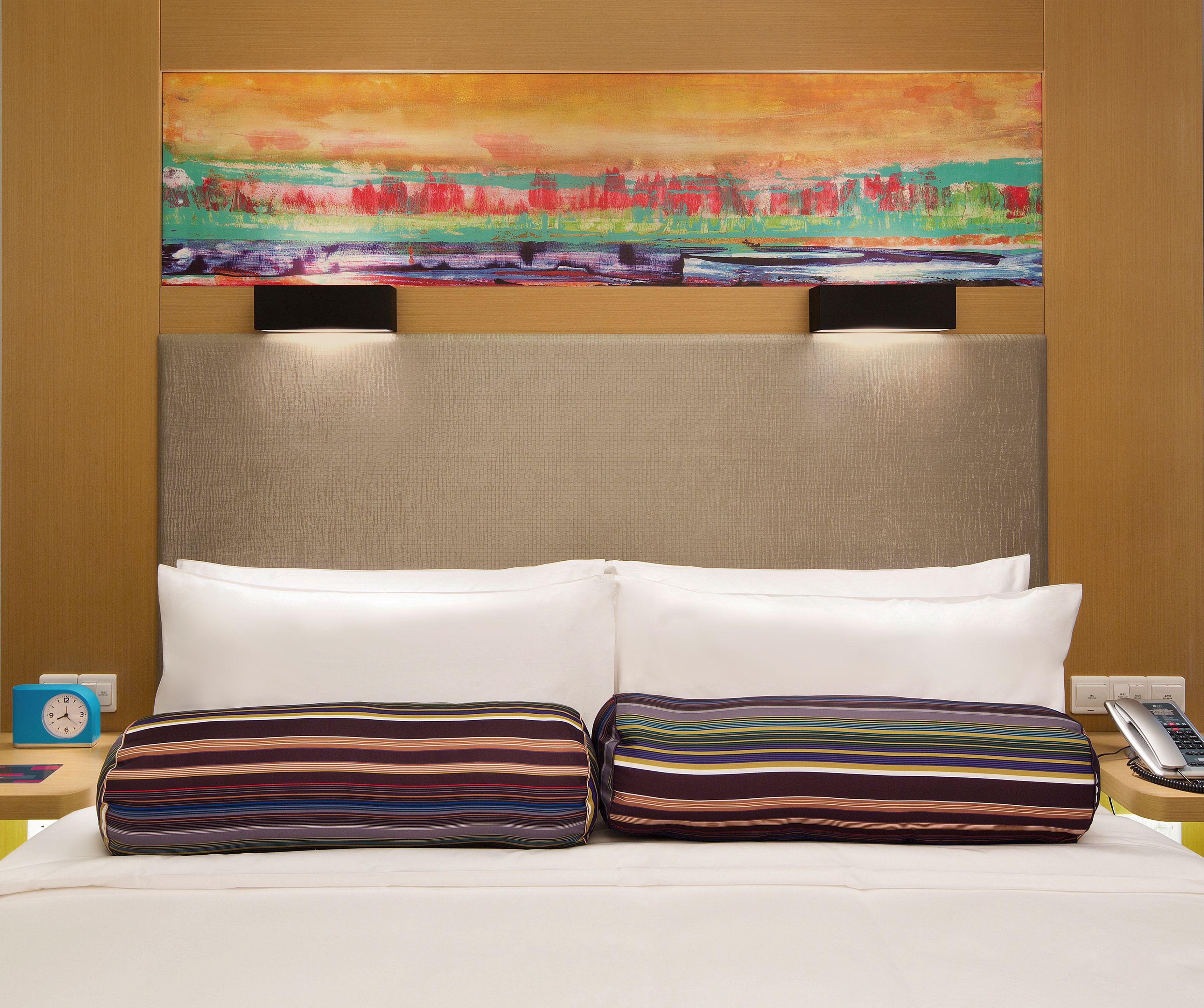 A hotel room with a large bed, two pillows, a painting above the bed, a nightstand with a clock, and a phone.