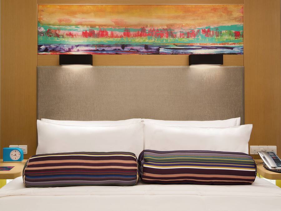 Une chambre d’hôtel avec un grand lit, deux oreillers, un tableau au-dessus du lit, une table de chevet avec une horloge et un téléphone.