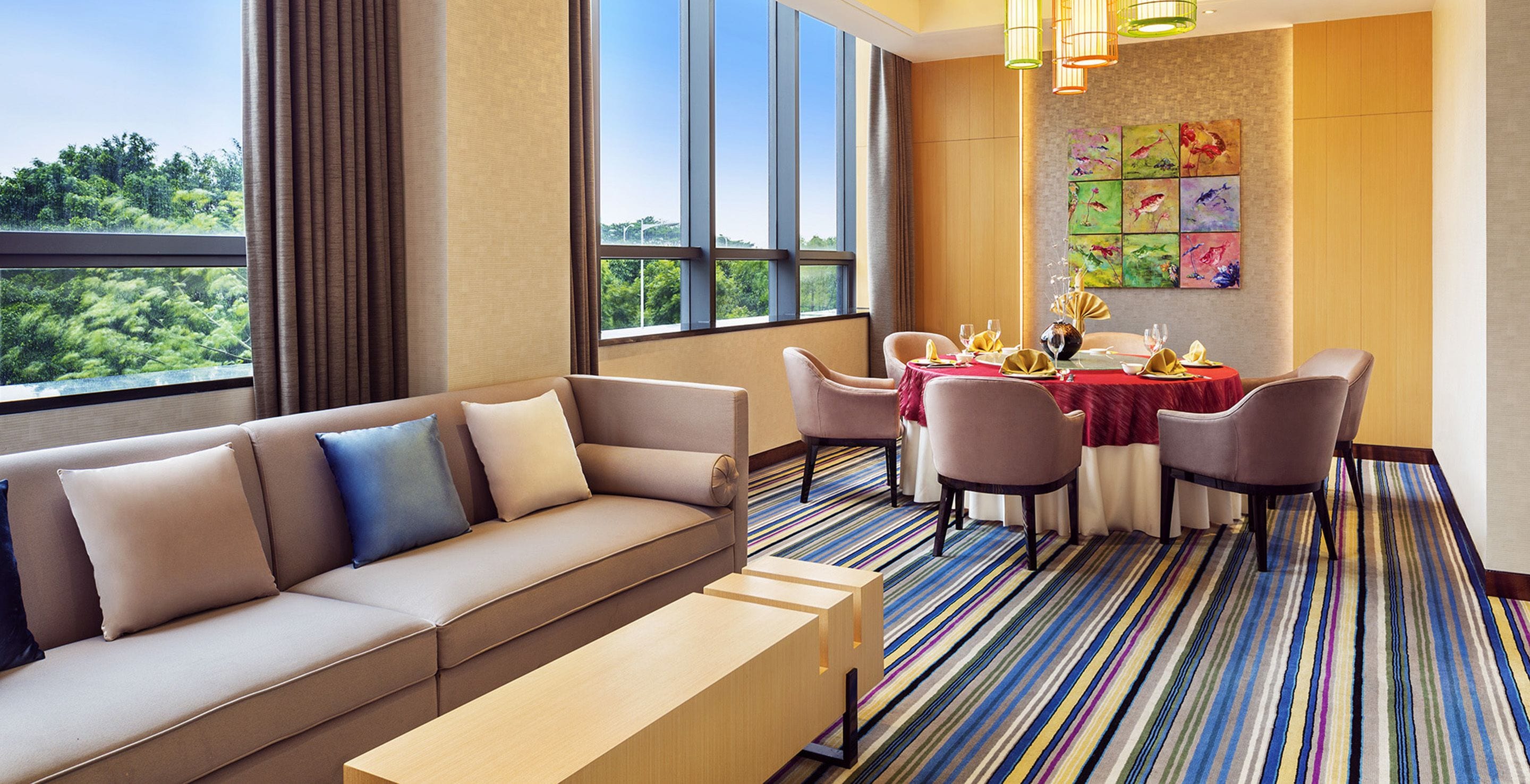 Una habitación de hotel moderna con sofá, mesa de comedor, sillas y vistas a zonas verdes a través de amplios ventanales.