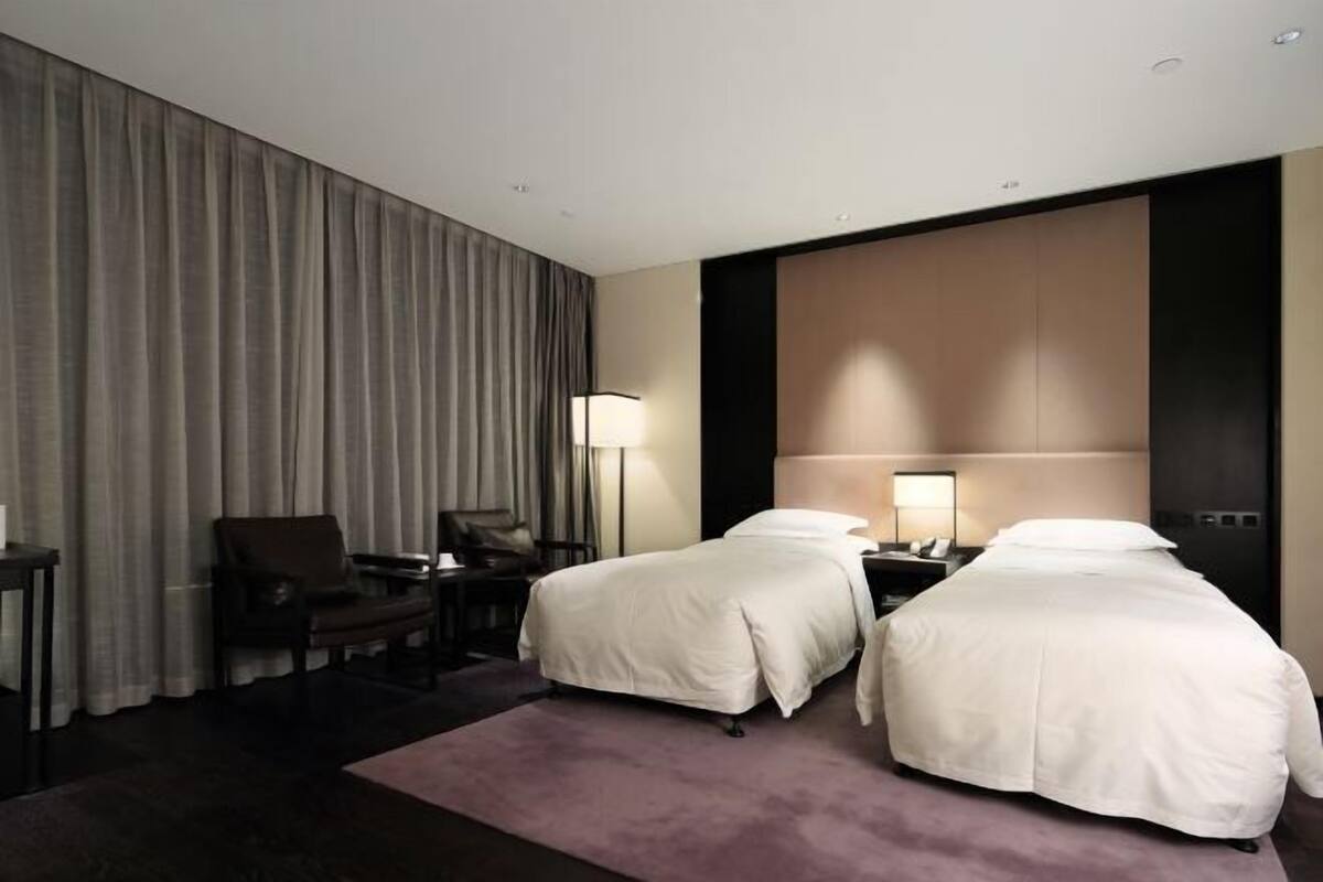 A hotel room with two beds, a desk, a chair, a lamp, and a large window with curtains.