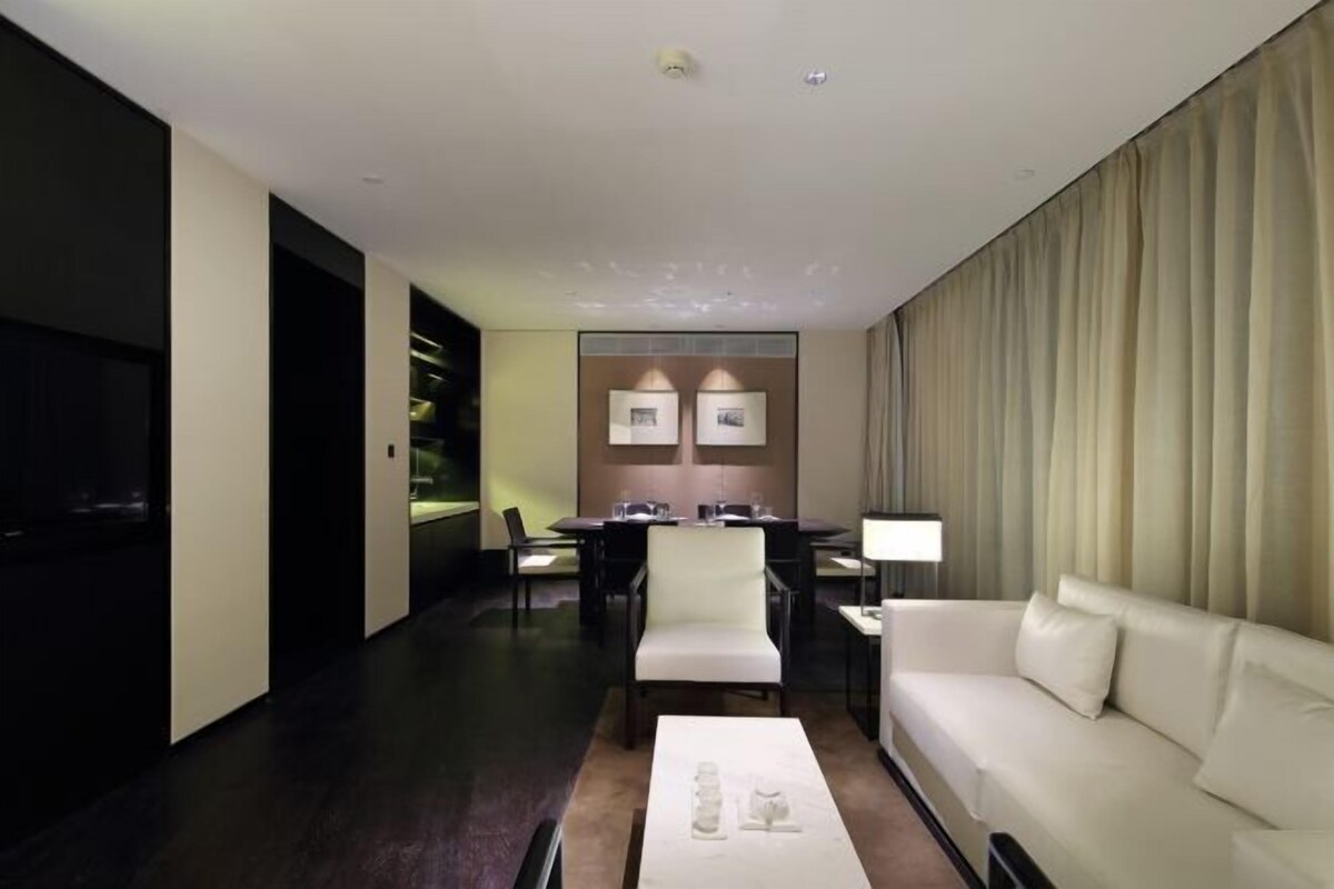 A modern hotel room with a dark wooden floor, a white sofa, a coffee table, a TV, and a dining area with chairs and a table.