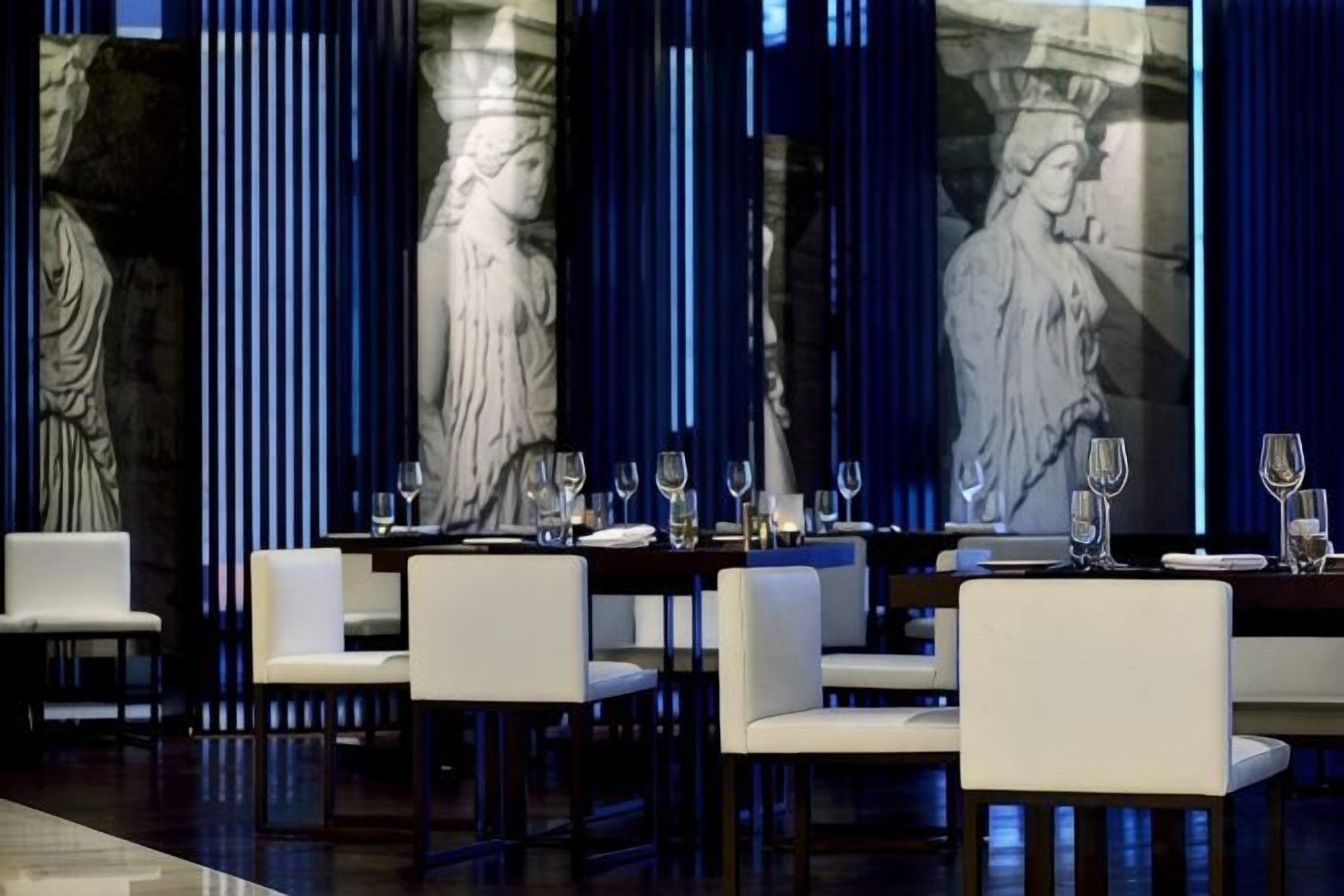 Un restaurant où les tables sont dressées pour le service, avec des colonnes en marbre et une grande fresque murale.