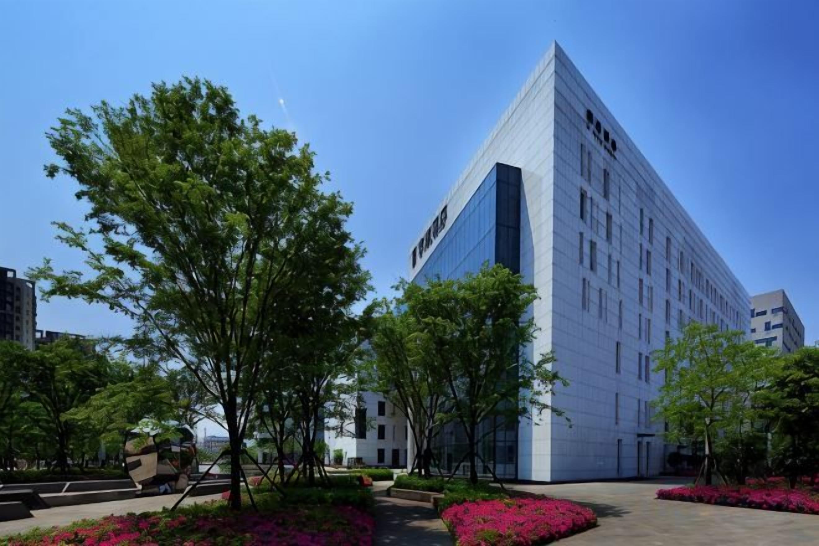 Een modern gebouw met een opvallende glazen gevel, omgeven door aangelegde gebieden met bomen en bloembedden.