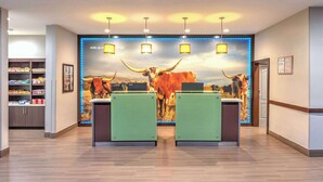 A reception area with a large bull painting, two green reception desks, overhead lighting, and a snack bar in the background. - La Quinta Inn & Suites by Wyndham Tyler - University Area (Tyler)