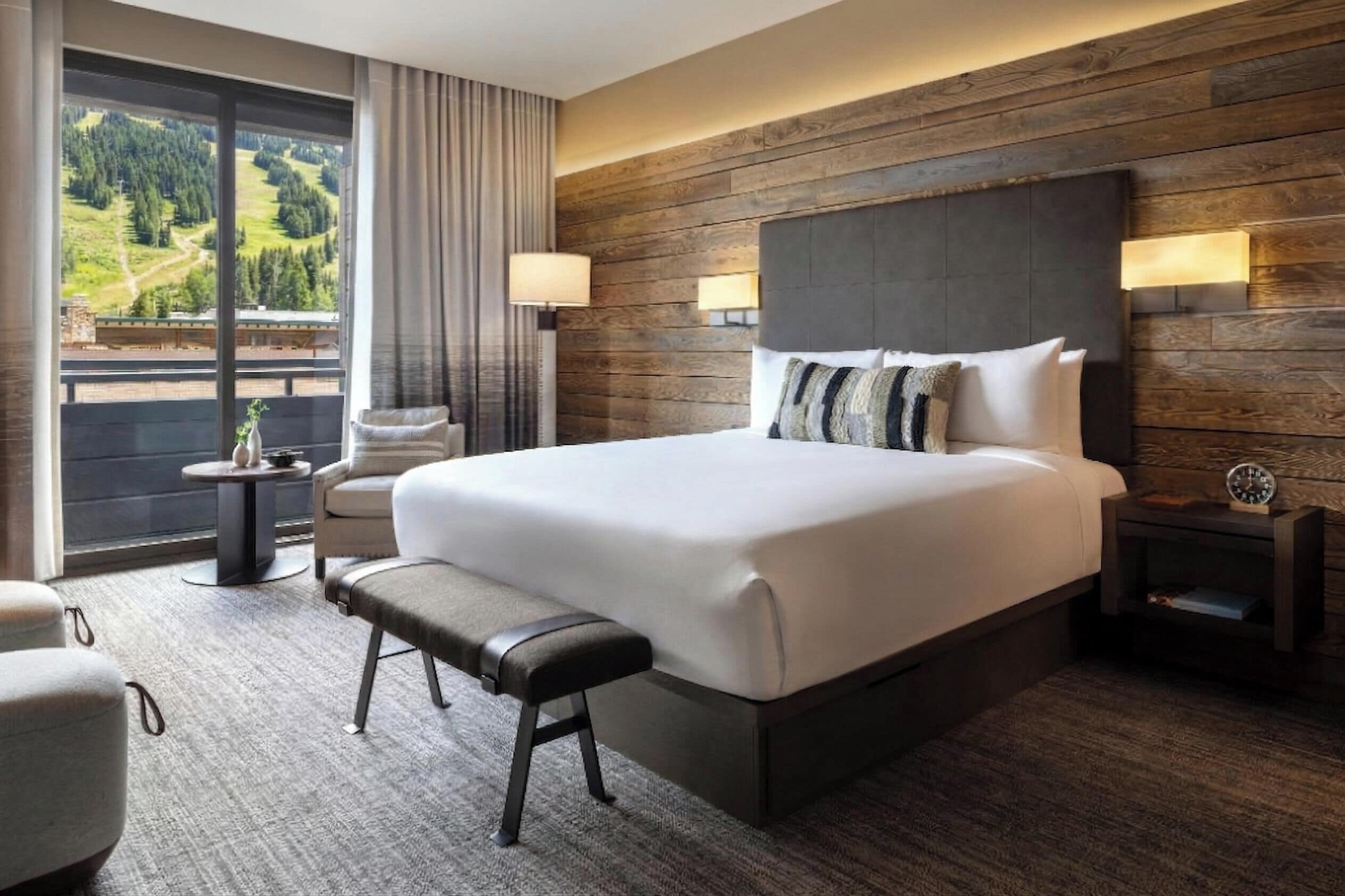 Una habitación de hotel moderna con una cama grande, una banca, una mesita pequeña y vistas a un paisaje verde a través de amplios ventanales.