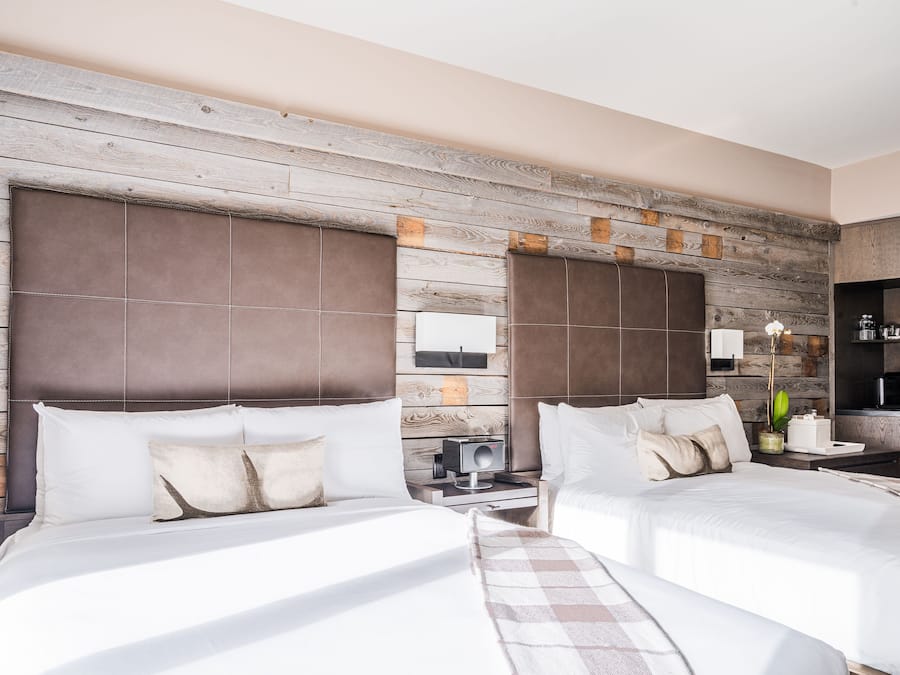 A hotel room with two beds, a wooden accent wall, a small bedside table with a lamp, and a coffee machine.